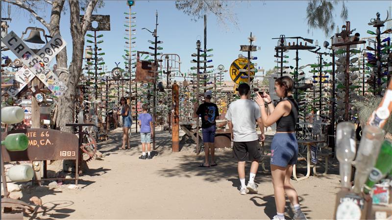 Visitors explore Elmer's Bottle Tree Ranch, a unique outdoor art installation featuring trees crafted from metal rods with colorful glass bottles. A video play button is also visible.